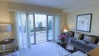 This single-floor residence offers a thoughtfully designed living space that balances comfort and style. The living room serves as the heart of the home, characterized by soft, neutral wall tones and plush carpeting that create a warm and inviting atmosphere. Large sliding glass doors flanked by sheer white floor-to-ceiling curtains flood the space with abundant natural light while framing a picturesque view of the surrounding outdoor area, including a pool and lush greenery. The seamless transition to a private balcony extends the living space outdoors, providing an ideal spot for relaxation and enjoying courtyard views. The living area is elegantly appointed with a contemporary gray sofa accented with decorative pillows, a sleek glass and metal coffee table, and tasteful décor including a coral accent piece and fresh floral arrangements. A side table with a soft-glowing lamp adds warmth to the space. The overall ambiance is light, airy, and refined, with a cohesive design palette that emphasizes clean lines and understated sophistication. The generous layout and bright, open feel make this a welcoming and functional living environment.
