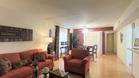 This single-floor space presents an open-concept layout that seamlessly connects the living and dining areas, creating a cohesive and functional flow throughout. The living area is anchored by rich, terracotta-toned upholstered seating, including a sofa and armchair, which complement the warm color palette of the space. A bold accent wall in deep red adds a striking visual element, contrasting effectively with the neutral cream-toned walls that surround it.

The flooring throughout is hardwood-style in a light natural finish, contributing to the bright and airy atmosphere of the space. A ceiling fan with an integrated light fixture is centrally positioned, providing both ventilation and ambient lighting. Natural light filters in through a large window dressed with patterned drapes, enhancing the overall warmth of the interior.

The dining area, visible beyond the living space, features dark wood dining chairs arranged around a table, with a tall potted plant nearby adding a touch of greenery. Decorative accents, including framed wall art and table accessories, are thoughtfully placed throughout, giving the space a well-curated, inviting character overall.