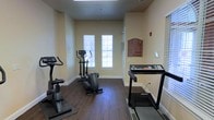 This fitness center offers a focused, functional workout environment designed to meet essential cardio needs. The room features warm beige walls and rich, dark hardwood-style flooring that lend a clean and inviting atmosphere. Natural light filters in through two windows fitted with white blinds, brightening the space and creating an open, airy feel.

The equipment layout is practical and well-organized, housing a treadmill along one wall, an elliptical trainer positioned centrally near the windows, and a stationary upright bike toward the front. This selection covers a range of low-impact cardiovascular workouts suited to a variety of fitness levels.

A posted fitness center rules sign near the door keeps the space orderly and ensures shared use remains respectful and safe. The room benefits from a straightforward, no-frills design that prioritizes usability. White baseboards and neutral wall tones contribute to the clean aesthetic throughout. A fire alarm is mounted on the wall, reflecting attention to safety standards. Overall, the space provides a convenient and accessible option for daily cardio exercise.