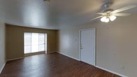 This single-floor space presents a straightforward, open layout anchored by rich, dark hardwood-style flooring that runs throughout the main living area. The room is defined by neutral tan walls and white trim, creating a clean, understated aesthetic. A ceiling fan with an integrated light fixture is centrally positioned, providing both air circulation and ambient lighting. Natural light enters through a large window fitted with horizontal blinds, illuminating the open space and highlighting the contrast between the warm flooring and the light-colored walls. The textured ceiling adds a subtle architectural detail overhead. A solid white entry door with brass hardware serves as the main access point, framed neatly against the surrounding walls. The space is open and unfurnished, allowing the full extent of the layout to be appreciated. White baseboards line the perimeter, providing a clean transition between the walls and flooring. Overall, the space offers a functional and practical environment with a neutral foundation that is well-suited to a variety of interior styles and furniture arrangements.