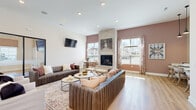 Located at 1564 McNaughten Rd in Columbus, Ohio, this single-floor clubhouse facility offers a thoughtfully designed, open-concept interior that blends functionality with modern style. The grand entrance greets visitors with soaring vaulted ceilings, floor-to-ceiling windows, and an abundance of natural light streaming through large glass doors, creating an immediately welcoming atmosphere. A professionally appointed reception desk sits just inside the entry, complemented by pendant lighting and a wall-mounted display screen. The expansive main gathering area features structural columns that define distinct zones without sacrificing the open feel. A central lounge is anchored by a stone-clad fireplace that stretches from floor to ceiling, flanked by large windows with views of the surrounding neighborhood. Rich hardwood-style flooring runs throughout, tying the spaces together cohesively. The kitchen is outfitted with white shaker cabinetry, white countertops, subway tile backsplash, stainless steel appliances, and a center island with bar seating beneath striking geometric pendant lights. A billiards table and additional seating areas round out the recreational offerings. Elegant brass chandeliers and recessed lighting add warmth, while a skylight in the central zone draws in additional daylight, making this a vibrant and versatile community gathering space.