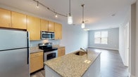 This single-floor residence offers a thoughtfully designed open-concept layout that seamlessly connects the kitchen, dining, and living areas. The kitchen is a standout feature, equipped with stainless steel appliances including a refrigerator, electric range with over-the-range microwave, and dishwasher. Light maple cabinetry extends to the ceiling for ample storage, while granite countertops carry through to the central island, which includes a built-in sink and is illuminated by stylish pendant lighting. Track lighting adds additional brightness to the cooking area. Durable wood-look flooring spans the main living spaces, creating a warm and cohesive aesthetic, while the open living and dining area benefits from natural light streaming through large windows fitted with blinds. The bedroom features plush carpet flooring and direct access to the outdoors through a glass door. The full bathroom includes a granite vanity with wood cabinetry, tile flooring, and a tiled tub-surround combination, while a second bathroom offers a granite vanity and accessible grab bar. A dedicated utility room houses a stacked washer and dryer alongside a water heater and electrical panel, providing practical in-unit convenience. Neutral gray walls throughout contribute to a clean, modern ambiance.