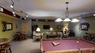 This single-floor entertainment space is thoughtfully designed for leisure and socializing. The open layout centers around a full-size pool table with a rose-felt surface, illuminated by an elegant multi-pendant light fixture that serves as a focal point overhead. Track lighting along the ceiling provides additional ambient coverage throughout the space.

The lounge area is warmly furnished with plush sofas and recliners, arranged around a wooden coffee table and accented by traditional table lamps that cast a cozy glow. A wall-mounted flat-screen television anchors the media portion of the room, making it well-suited for both active game play and relaxed viewing.

The walls are finished in a muted olive green with a contrasting dark brown accent band near the ceiling, lending the space a rustic, cabin-inspired character. Neutral carpeting runs throughout, adding comfort underfoot. A bar-height table with stools is tucked along one side, offering a casual spot for dining or gathering. A wooden storage cabinet and framed artwork on the walls complete the décor. A private door in the corner leads to an additional room, adding functional versatility to this well-appointed entertainment retreat.
