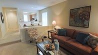 This single-floor residence offers an inviting and well-appointed layout designed for comfortable everyday living. The living area serves as the heart of the home, featuring warm neutral wall tones, carpeted flooring, and ample space for relaxed seating arrangements. A plush sofa and accent chair provide comfortable furnishing options, complemented by a glass-top coffee table and coordinating side table with a classic table lamp that adds a soft, warm glow to the room. A half-wall partition elegantly separates the living space from the kitchen, creating an open yet defined flow between the two areas. The kitchen is equipped with cabinetry in a warm wood finish, a countertop breakfast bar with seating, and a window that allows natural light to brighten the space. Recessed lighting and strategically placed windows throughout contribute to the overall sense of warmth and openness. The neutral color palette throughout the space creates a calm, cohesive ambiance that is both functional and welcoming. The thoughtful layout maximizes the use of space, making this residence an efficient and comfortable place to call home.
