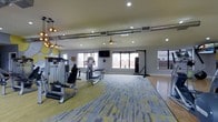 This single-floor fitness center is designed to support a wide range of workout routines in a bright, open environment. The space is thoughtfully divided into distinct training zones: one side features strength training machines, including a cable machine and a seated press station, arranged on patterned carpet tiles in a striking yellow and gray palette. The other side transitions to a smooth hardwood floor, offering open space ideal for stretching, yoga, or bodyweight exercises, and is complemented by a freestanding heavy bag for boxing and martial arts training. Natural light pours in through multiple large windows that line the perimeter walls, creating an airy and energizing atmosphere. Overhead, exposed silver ductwork runs across the ceiling, adding an industrial design element that contrasts with the otherwise clean, modern interior. Ceiling fans and recessed lighting ensure the space remains comfortable and well-lit throughout the day. A wall-mounted flat-screen TV is positioned for easy viewing during workouts. Decorative pendant lighting near the entrance adds a polished, welcoming touch. The overall design balances functionality with contemporary aesthetics, making this fitness center both practical and visually appealing.