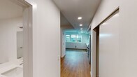 This single-floor unit showcases a clean, modern aesthetic defined by crisp white walls, warm hardwood flooring, and recessed lighting throughout. A central hallway connects the main living areas, leading from the entry toward a bright, windowed living space at the far end. Natural light filters in through large windows, lending the open living area an airy, welcoming quality.

The kitchen is a standout feature, designed with a contemporary sensibility. High-gloss, floor-to-ceiling cabinetry in soft grey and white tones provides ample storage, while a continuous white countertop runs the length of the workspace. Stainless steel appliances include a full-size refrigerator, a professional-style range with a built-in microwave above, and an integrated dishwasher, all contributing to a cohesive, polished look. White tile backsplash adds a subtle texture against the cabinetry.

The living area benefits from an open layout with windows that bring in natural light, while a closeted storage area provides practical organization. The bathroom maintains the unit's minimalist theme, featuring a long white vanity with an integrated sink, a wall-mounted mirror, wood-tone flooring, and a tiled shower area. Recessed lighting and white walls keep the space feeling clean and bright throughout.