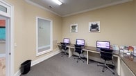 This single-floor space offers a polished and functional environment designed to accommodate both welcoming and productive activities. The entryway sets an inviting tone, featuring a glass-paneled door with a transom window above, light hardwood flooring, and a decorative round table centered on a vibrant patterned area rug — creating an elegant first impression. Warm beige walls and white trim carry throughout the space, lending a cohesive and professional aesthetic. The business center is the focal point of the interior, thoughtfully appointed with a long granite-topped desk that runs the length of the room, accommodating multiple computer workstations and a multifunction printer. Ergonomic task chairs provide comfortable seating for extended use. Crown molding and recessed lighting enhance the room's refined character, while wall-mounted artwork featuring vintage typewriter prints adds a creative, story-driven touch. Glass-paneled interior doors connect the rooms, maintaining an open and accessible flow. Plush carpeting in the workstation area provides a quiet, comfortable atmosphere, making this space well-suited for focused work and seamless connectivity.