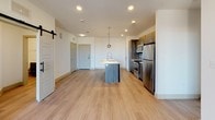 This single-floor residence showcases a contemporary design with a thoughtful layout visible in its open floor plan. The kitchen serves as a central highlight, featuring dark wood-grain cabinetry extending to the ceiling, a full suite of stainless steel appliances including a range, microwave, and refrigerator, and a large island with a white quartz countertop, undermount sink, and pendant lighting overhead. Light wood-tone flooring flows throughout the main living and kitchen areas, complementing the clean white walls and recessed lighting. A sliding barn door adds a stylish architectural accent near the living space, which also offers access to an outdoor area through a glass-paneled door.

The bedrooms are finished with plush carpeting, ceiling fans with light fixtures, and windows fitted with horizontal blinds that allow natural light while maintaining privacy. The primary bathroom is particularly well-appointed, offering dual backlit vanity mirrors, a double-sink quartz countertop with dark wood cabinetry below, wood-look tile flooring, large-format gray wall tiles, a glass-enclosed walk-in shower with a rain showerhead, and a soaking tub. The overall aesthetic throughout is clean, modern, and cohesive.