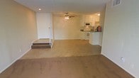 This single-floor space features an open-concept layout that connects the main living area to the kitchen. The living area is carpeted in a neutral beige tone, providing a comfortable and understated foundation. A ceiling fan with a light fixture is centrally positioned overhead, offering both airflow and ambient lighting. A front entry door is accessible via a short set of steps that lead down into the main living space, creating a subtle split-level effect.

The kitchen is situated toward the rear of the open area and features white cabinetry, white appliances including a range and microwave, and a darker countertop that provides contrast against the lighter cabinetry. The kitchen transitions smoothly from the carpeted living space via a tile or hard surface floor, visually defining the functional cooking area. A small peninsula or counter divider separates the kitchen from the living space, offering a natural boundary between the two areas. The overall color palette throughout the space is light and neutral, with white walls contributing to a clean, open atmosphere.