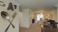 This single-floor residence offers a thoughtfully designed open layout that connects key living areas in a seamless and functional flow. The hallway serves as the central spine of the home, lined with white paneled doors that provide access to individual rooms, and accented with crown molding that lends a classic, refined touch to the interiors throughout.

The living area is bright and inviting, benefiting from a large window dressed with vertical blinds that allows generous natural light to fill the space. A ceiling fan with an integrated light fixture anchors the room, offering both comfort and practicality. Plush wall-to-wall carpeting runs through the living and hallway areas, adding warmth and softness underfoot.

Adjacent to the main corridor, a dining area is visible, furnished with a dark wood table and chairs, creating a defined yet open dining space. Decorative wall art — including oversized utensil-style pieces — adds personality and character to the entryway walls.

The overall ambiance is warm, neutral, and comfortable, with cream and beige tones dominating the walls and flooring. The layout is efficiently organized, making excellent use of the available space while maintaining a spacious and uncluttered feel throughout.