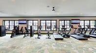 The Kempton Luxury Living fitness center at 425 Devon Bryan St. in Columbus, Ohio is a generously sized, single-floor gym facility designed to deliver a premium workout experience. The space is anchored by a welcoming reception area featuring a sleek, dark wood credenza with brass hardware, a large branded display, and tasteful décor — setting an elevated tone from the moment you arrive.

Throughout the gym, a distinctive patterned carpet in soft greens and neutrals adds a refined aesthetic underfoot, while white walls with rich green crown molding and baseboards reinforce the community's signature color palette. Recessed lighting and ceiling fans with pendant mounts ensure the space remains bright and comfortable during any workout.

Large, dark-framed windows line multiple walls, flooding the interior with natural light and offering views of the surrounding neighborhood. The fitness floor is comprehensively equipped, featuring a full row of treadmills and ellipticals with integrated screens, spin bikes, a multi-station cable and strength machine circuit, free weights, medicine balls, and dedicated stretching zones. Full-length mirrors on select walls enhance the sense of openness and support proper form during training.

Branded "Kempton Luxury Living" signage is displayed prominently throughout, reinforcing the community identity within a space that balances functionality with upscale design.