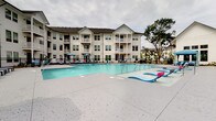 Located at 1001 Linger Longer Dr in Summerville, South Carolina, this outdoor amenity area is centered around a generously sized swimming pool with crystal-clear blue water and a wide, textured concrete pool deck. The deck is well-appointed with chaise lounge chairs, side tables, and colorful umbrellas, providing ample space for relaxing in the sun. A covered cabana-style structure with white columns offers shaded seating areas directly adjacent to the pool, ideal for those seeking relief from the South Carolina heat. A separate covered porch area features classic white rocking chairs arranged along white shiplap-clad walls, creating an inviting Southern-style retreat with views of the pool courtyard. The surrounding landscape includes mature palm trees, tall pines, and neatly mulched planting beds, lending a lush, resort-like atmosphere to the space. A black wrought iron security fence with a gated entrance encloses the pool area, ensuring privacy and safety. The multi-story residential buildings surrounding the courtyard feature private balconies that overlook the pool, reinforcing the community-centered design of this outdoor space. The overall ambiance is bright, open, and welcoming, with a well-maintained aesthetic throughout.