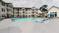 Located at 1001 Linger Longer Dr in Summerville, South Carolina, this outdoor amenity area is centered around a generously sized swimming pool with crystal-clear blue water and a wide, textured concrete pool deck. The deck is well-appointed with chaise lounge chairs, side tables, and colorful umbrellas, providing ample space for relaxing in the sun. A covered cabana-style structure with white columns offers shaded seating areas directly adjacent to the pool, ideal for those seeking relief from the South Carolina heat. A separate covered porch area features classic white rocking chairs arranged along white shiplap-clad walls, creating an inviting Southern-style retreat with views of the pool courtyard. The surrounding landscape includes mature palm trees, tall pines, and neatly mulched planting beds, lending a lush, resort-like atmosphere to the space. A black wrought iron security fence with a gated entrance encloses the pool area, ensuring privacy and safety. The multi-story residential buildings surrounding the courtyard feature private balconies that overlook the pool, reinforcing the community-centered design of this outdoor space. The overall ambiance is bright, open, and welcoming, with a well-maintained aesthetic throughout.