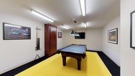 This single-floor game room is thoughtfully designed as a dedicated recreational space, offering an engaging environment for leisure and entertainment. At the heart of the room sits a full-size pool table with a blue felt surface and a dark wood frame, positioned beneath a stylish multi-shade pendant light that provides focused illumination for gameplay. The bold yellow floor area beneath the table creates a striking visual contrast against the darker border flooring, giving the space a lively, dynamic character. A wall-mounted cue rack keeps several pool cues neatly organized and within easy reach, while a wooden storage cabinet offers additional functionality. Framed artwork and posters adorn the white walls, adding personality and a sense of casual charm. A flat-screen television is mounted on the wall opposite the table, making the room suitable for watching sports or entertainment between games. Overhead fluorescent lighting ensures the entire space is well-lit, complementing the pendant fixture above the table. The overall ambiance is energetic yet functional, striking a balance between a casual rec room and a well-equipped entertainment space ideal for socializing and friendly competition.