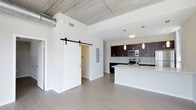 This single-story unit showcases a thoughtfully designed open-concept layout that seamlessly connects the living area and kitchen. An exposed concrete ceiling with visible ductwork adds an industrial character, complemented by crisp white walls and smooth light-toned hardwood-style flooring throughout. A striking black sliding barn door serves as a functional and stylish architectural accent, providing access to an adjoining room while enhancing the modern aesthetic.

The kitchen is a standout feature, equipped with dark espresso cabinetry, a subway tile backsplash, and stainless steel appliances including a microwave and range. A central island with a white countertop and undermount sink offers additional workspace and casual seating potential. Pendant lighting fixtures hang above the island, providing both task lighting and visual interest.

The open living space benefits from a bright, airy feel thanks to the high ceilings and clean, minimalist design. The combination of industrial and contemporary elements throughout gives the unit a distinctive, well-balanced character suitable for modern living.