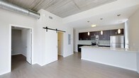 This single-story unit showcases a thoughtfully designed open-concept layout that seamlessly connects the living area and kitchen. An exposed concrete ceiling with visible ductwork adds an industrial character, complemented by crisp white walls and smooth light-toned hardwood-style flooring throughout. A striking black sliding barn door serves as a functional and stylish architectural accent, providing access to an adjoining room while enhancing the modern aesthetic.

The kitchen is a standout feature, equipped with dark espresso cabinetry, a subway tile backsplash, and stainless steel appliances including a microwave and range. A central island with a white countertop and undermount sink offers additional workspace and casual seating potential. Pendant lighting fixtures hang above the island, providing both task lighting and visual interest.

The open living space benefits from a bright, airy feel thanks to the high ceilings and clean, minimalist design. The combination of industrial and contemporary elements throughout gives the unit a distinctive, well-balanced character suitable for modern living.