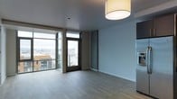 This single-floor space showcases a sleek, contemporary design with an open and airy feel. The living area is anchored by expansive floor-to-ceiling glass windows and a glass-paneled balcony door, flooding the interior with abundant natural light while offering sweeping urban views of the surrounding cityscape. The balcony access adds an attractive outdoor extension to the living space.

Light-toned hardwood flooring runs throughout, contributing to the bright and open atmosphere and complementing the neutral color palette. Smooth, flat ceilings are fitted with a simple flush-mount drum light fixture, maintaining the clean, minimalist aesthetic. The walls are finished in a soft, cool gray tone that pairs well with the modern architectural elements.

Structural columns with warm wood-toned cladding provide visual contrast against the cooler gray tones, adding depth to the space. A stainless steel appliance is partially visible, hinting at a kitchen area nearby. The overall ambiance is modern, understated, and functional, with an emphasis on natural light, urban connectivity, and clean architectural lines throughout.