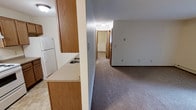 This single-floor unit offers a practical, efficient layout designed for everyday living. The kitchen is positioned at the entrance and features warm wood-toned cabinetry, a double-basin sink set into a light-colored countertop, and a white refrigerator, providing ample storage and workspace. Tile flooring anchors the kitchen area, transitioning to neutral wall-to-wall carpeting that extends throughout the living space and hallway beyond.

A half-wall partition separates the kitchen from the adjoining carpeted living area, creating a sense of division while maintaining an open feel between the two spaces. The living area is generously sized with light gray walls and flush-mount ceiling lighting that provides even illumination throughout.

A central hallway extends from the main living area toward the rear of the unit, lined with wood-toned doors that lead to additional rooms, including what appears to be a bathroom and one or more bedrooms. The hallway features a baseboard heater, indicating a practical heating setup throughout. The overall ambiance is clean, neutral, and move-in ready, suited for straightforward, comfortable living.