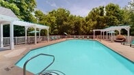 Located at 405 Thomas Burke Dr in Hillsborough, NC, this outdoor amenity area centers around a generously sized swimming pool with a classic geometric shape and clearly marked depth indicators. The pool deck is finished in a stamped concrete surface that wraps around the entire perimeter, providing ample space for relaxation and recreation. Teal lounge chairs are arranged along the poolside, offering comfortable spots for sunbathing, while white patio tables paired with chairs and teal umbrellas provide shaded seating areas perfect for leisure. Two white pergolas flank the pool area, adding architectural character and additional covered retreats. A brick building adjacent to the pool serves as a functional support structure, likely housing restroom or storage facilities. The entire area is enclosed by a white railing fence, ensuring safety while maintaining an open, airy feel. Mature, leafy trees form a natural green backdrop on all sides, creating a sense of privacy and a peaceful, park-like atmosphere. The combination of well-maintained landscaping, coordinated furnishings, and thoughtful layout makes this outdoor space an inviting destination for warm-weather enjoyment.