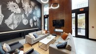 This community room is a thoughtfully designed shared space that combines sophisticated aesthetics with practical amenities across a single floor. The lounge area commands attention with its soaring ceilings and a dramatic floor-to-ceiling wood-paneled wall, which anchors a sleek wall-mounted television and a linear fireplace below. Comfortable gray sofas arranged around a minimalist white coffee table create an inviting gathering area, accented by warm-toned throw pillows and a natural fiber area rug. Circular pendant light fixtures provide ambient illumination overhead, while large windows invite natural light and offer views of the exterior surroundings. A bold, large-scale botanical mural rendered in monochromatic tones covers an entire accent wall, serving as a striking artistic focal point. The fully equipped kitchen features professional-grade stainless steel appliances, including double stacked wall ovens and a full-size refrigerator, complemented by white quartz countertops, a subway tile backsplash, warm wood cabinetry, and polished concrete flooring. This kitchen is clearly suited for community events and gatherings. Together, these elements create a refined yet welcoming environment ideal for social engagement.