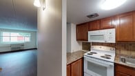 This single-floor unit offers a straightforward, functional layout with consistent dark hardwood-style flooring running throughout all living areas, bedrooms, and the bathroom, complemented by white walls and dark baseboards that create a clean, cohesive aesthetic.

The kitchen serves as a practical hub of the unit, featuring warm-toned medium wood cabinetry both above and below the counters, a stone-patterned laminate countertop, and a stainless steel sink. A tile mosaic backsplash in neutral brown tones adds visual texture behind the white electric coil range and over-the-range microwave. The kitchen opens toward the main living area, which features an open-concept flow with a peninsula-style counter dividing the two spaces.

The living area is generously proportioned, offering an open view toward the kitchen and access to the rest of the unit via a central hallway. Multiple rooms, likely serving as bedrooms, each feature dark hardwood-style flooring, large windows that bring in natural light and outdoor views, textured ceiling tiles, and wall-mounted climate control units. The rooms are neutral and move-in ready.

The bathroom includes a full-size soaking tub and shower combination with clean white subway tile surround, standard fixtures, and dark hardwood-style flooring, maintaining the unit's consistent flooring theme throughout.
