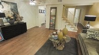 This two-story space offers an open and airy living area anchored by rich hardwood flooring that extends throughout the main level. The living room draws immediate attention with a distinctive round glass-top coffee table supported by a sculptural driftwood base, paired with a dark area rug that grounds the seating area. A ceiling fan with integrated lighting overhead provides both comfort and illumination. A tall, rustic mirror in a dark wood frame serves as a bold decorative focal point along one wall, while a collection of framed artwork adds a personal touch nearby. Built-in storage and closet space contribute to the room's functionality, while baseboard heating ensures year-round comfort. A white-railed staircase with carpeted treads leads to the upper level, offering visual connectivity between floors and adding architectural interest to the space. The overall ambiance is warm and eclectic, blending natural materials like wood and stone with clean, neutral walls that allow the decor to take center stage. The layout feels spacious and well-proportioned, providing a comfortable and inviting environment throughout.