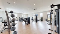 Located in Fairhope, Alabama, this well-appointed fitness center offers a generous, open-plan exercise space designed to accommodate a wide range of workout routines. The room features light hardwood-style flooring, neutral walls, and recessed lighting throughout, creating a bright and clean environment conducive to focused training. Ceiling fans are positioned throughout the space to maintain comfortable airflow.

The facility is comprehensively equipped with a full suite of TRUE-brand strength machines, including cable systems, chest press, leg press, and multi-functional units. A dedicated free weights area features a full dumbbell rack and medicine ball storage, complemented by a large wall-mounted mirror that enhances the sense of space. The cardio section lines one windowed wall and includes multiple treadmills and elliptical trainers, all facing large windows that provide natural light and views of the surrounding community and outdoor amenity areas. Wall-mounted flat-screen televisions are positioned throughout for entertainment during workouts.

Weight storage, plate-loaded benches, and open floor space allow for versatile training options. The layout offers ample room between stations, ensuring comfort and safety. Direct exterior access is available through a glass-paned door, seamlessly connecting the fitness center to the outdoor surroundings.