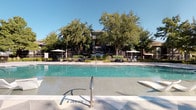 Located at 3501 W Walnut Hill Ln in Irving, Texas, this amenity space showcases a thoughtfully designed fitness center and a resort-style outdoor pool area. The fitness center is highlighted by dramatic vaulted ceilings adorned with bold black exposed beams, creating an open and energizing atmosphere. Large black-framed windows flood the space with natural light while offering views of the surrounding landscape. The gym is well-appointed with a comprehensive selection of equipment, including weight machines, a cable/functional trainer system, free weights with a dedicated rack, ellipticals, and cardio machines. Full-length mirrors and motivational wall art contribute to an engaging workout environment, while striped carpet flooring adds a modern aesthetic touch. A granite-topped counter station provides additional utility within the space. The outdoor pool area presents a generously sized swimming pool with clean, light-tiled surroundings. Sleek lounge chairs are positioned poolside, and multiple large patio umbrellas offer shaded seating areas. The pool deck is framed by mature trees that provide a lush, park-like setting, with the residential buildings visible in the background. Together, these amenities deliver a well-rounded recreational experience for residents.