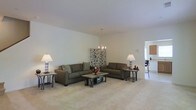 This two-story home offers a well-designed layout that balances openness with functional living. The main living area is generously proportioned, featuring plush wall-to-wall carpeting in a warm neutral tone that complements the crisp white walls and high ceilings. An elegant pendant chandelier serves as a focal point overhead, adding a refined touch to the space. The room comfortably accommodates a full seating arrangement, making it ideal for both everyday living and entertaining. Ambient floor lamps on either side of the seating area contribute to the warm, inviting atmosphere. A decorative room divider panel adds an artistic accent to the living space. The open sightline connects the living area directly to the kitchen, which is visible in the background and features cabinetry and bar-style seating, enhancing the home's overall sense of connectivity. Natural light filters into the space, reinforcing the bright, airy ambiance throughout. The two-floor configuration provides additional living space, offering versatility and room to accommodate a variety of needs. The overall design aesthetic is clean, modern, and understated, making it a welcoming and adaptable environment.