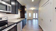 This thoughtfully designed studio presents an efficient, single-floor open layout that maximizes every square foot. The kitchen is a standout feature, showcasing rich dark cabinetry that extends from floor to ceiling, offering generous storage capacity. A speckled granite-style countertop with an undermount sink complements the sleek stainless steel appliances, including a dishwasher and range, creating a polished, contemporary aesthetic. The cabinetry's deep tones contrast beautifully against the light-colored walls, lending the space a bold yet balanced visual appeal. Wide-plank hardwood-style flooring runs continuously throughout, unifying the kitchen and living areas with warmth and cohesion. The open living area benefits from abundant natural light, courtesy of a set of elegant French doors flanked by sidelights at the far end of the space. Recessed and flush-mount ceiling lighting ensures the interior remains bright and welcoming at any hour. Clean white trim and baseboards frame the space neatly, while a secondary door offers additional access or closet space. The overall ambiance is modern and move-in ready, blending functional design with contemporary finishes.