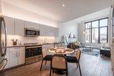 This single-floor residence showcases a thoughtfully designed open-concept layout that seamlessly connects the kitchen, dining, and living areas. The kitchen is outfitted with sleek, light blue flat-front cabinetry extending to the ceiling, under-cabinet lighting, a white subway tile backsplash, stainless steel appliances including a range, built-in microwave, dishwasher, and refrigerator, and a undermount sink with a modern pull-down faucet. Light wood-style flooring runs throughout the main living areas, contributing to a cohesive and airy aesthetic.

The open living area benefits from impressively tall ceilings and large, black-framed floor-to-ceiling windows that frame expansive urban skyline views and flood the space with natural light. The bedroom continues this design language, featuring the same striking oversized windows with panoramic city views and elevated ceilings that enhance the sense of volume.

The bathroom is finished in clean white rectangular subway tile surrounding a full soaking tub and shower combination, maintaining the minimalist aesthetic found throughout. Overall, the space presents a modern, urban character with a bright, neutral palette, high-end finishes, and an emphasis on natural light and city-facing views.