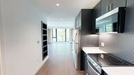 Located at 31 Artful Way in Asheville, NC, this contemporary single-floor studio offers a well-planned layout that maximizes every square foot. The open-concept kitchen and living area forms the heart of the space, seamlessly connecting daily living and cooking functions. The kitchen is outfitted with sleek, dark cabinetry that extends floor-to-ceiling, stainless steel appliances including a full-size refrigerator, electric range, and microwave, complemented by white quartz countertops and a geometric hexagonal tile backsplash that adds a modern touch. Wood-tone flooring runs throughout, lending warmth and continuity to the interior. The living area is bright and airy, anchored by large, black-framed floor-to-ceiling windows that invite abundant natural light and offer views of surrounding greenery and the Asheville cityscape. Neutral-toned draping panels frame the windows, adding softness to the clean-lined design. Recessed lighting throughout maintains a polished, uncluttered ceiling plane. Two closets provide practical storage solutions. The bathroom features a minimalist aesthetic with a floating vanity, black hardware accents, light tile flooring, and a narrow window that brings in natural light while maintaining privacy. Overall, the space presents a refined, modern ambiance suited to contemporary urban living in the heart of Asheville.