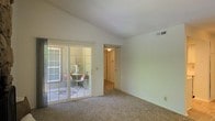 This single-floor residential space offers a comfortable and functional layout with a clean, neutral aesthetic throughout. The main living area is finished with wall-to-wall carpeting that adds warmth underfoot, while smooth white walls and a vaulted ceiling create a sense of openness and airiness. A standout feature of the living space is the large sliding glass door framed with white trim, which floods the interior with natural light and provides direct access to a covered outdoor patio. The patio offers a cozy retreat, visible through the glass and accented by decorative furnishings. A ceiling-mounted light fixture in the hallway beyond the main room provides gentle ambient illumination, and a connecting hallway leads further into the home, suggesting additional rooms beyond. Overhead HVAC vents are neatly integrated into the ceiling, contributing to the tidy, well-maintained appearance of the space. The overall ambiance is bright, calm, and inviting, with a simple design that offers a versatile canvas for personalization. The seamless connection between the indoor living area and the outdoor patio enhances the sense of space and makes this a particularly appealing feature of the home.