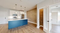 Located at 300 YMCA Way in Penfield, NY, this thoughtfully designed 2-bedroom, 1-bathroom residence offers a well-appointed single-floor layout with contemporary finishes throughout. Light-toned vinyl plank flooring runs continuously through every room, creating a cohesive and clean aesthetic.

The kitchen serves as the centerpiece of the home, anchored by a striking navy blue island topped with white quartz countertops and an undermount sink. White shaker-style cabinetry with matte black hardware lines the perimeter, complemented by a white tile backsplash and a full suite of stainless steel appliances including a range, over-the-range microwave, dishwasher, and refrigerator. Three pendant lights hang above the island, adding warm, focused illumination. A bold natural wood-plank accent wall borders the kitchen and dining area, providing a striking textural contrast.

The living room features large windows and a glass door that invite abundant natural light and offer views of the surrounding green landscape. Both bedrooms are generously proportioned, each with windows overlooking the outdoors. The primary bedroom includes a walk-in closet accessible through a paneled white door. The bathroom is finished with a white vanity, quartz countertop, matte black fixtures, and matching wood-look flooring.