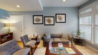 This single-story space is thoughtfully designed with multiple distinct functional areas unified by a consistent palette of soft blue-gray walls, white trim, and warm hardwood and tile flooring throughout. The central great room is a standout feature, boasting soaring vaulted ceilings that amplify the sense of openness. A classically detailed white fireplace surround with granite accents anchors one wall, complemented by a wall-mounted television above. Adjacent to the great room, a generous dining nook is framed by a full bay of white plantation-shuttered windows, flooding the area with natural light and offering views of surrounding greenery. A round glass-top dining table occupies this bright corner comfortably. The open kitchen features white cabinetry, tile flooring, and standard appliances, with easy flow into the main living area. A dedicated fitness space provides room for exercise equipment, while a welcoming reception lounge area includes tufted upholstered seating, a glass coffee table, framed floor plan artwork, and plantation-shuttered windows. The overall ambiance is clean, versatile, and well-lit, with a cohesive design that balances comfort and functionality across each zone.