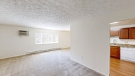 This single-floor residence offers a straightforward and functional layout encompassing two bedrooms, one bathroom, a living room, kitchen, and hallway. Throughout the unit, neutral beige carpeting and cream-colored walls create a consistently light and clean aesthetic, while textured ceilings add a subtle decorative detail visible across multiple rooms.

The living room is a generously sized space featuring a large window fitted with vertical blinds, allowing ample natural light. Baseboard heating runs along the walls, and a wall-mounted air conditioning unit provides additional climate control. The open feel of the room makes it well-suited for a variety of furniture arrangements.

The kitchen is equipped with warm-toned wood cabinetry, stone-look countertops, and a white electric range with an overhead exhaust hood, providing practical functionality with a cohesive design.

The spacious primary bedroom features its own window with vertical blinds, baseboard heating, and direct access to the full bathroom, which features tile flooring. A closet with a wood-paneled door provides dedicated storage. The second bedroom is similarly carpeted with a window and baseboard heater. A hallway connects the main living areas to the bedrooms and bathroom.