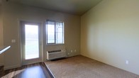 This single-floor space features a straightforward, functional layout with a blend of hard flooring and carpeting. The main living area is finished with beige carpet, while the entryway transitions to smooth hard flooring, creating a practical delineation between zones. Neutral-toned walls throughout lend a clean, understated aesthetic that allows for flexible interior styling.

Natural light enters through a window fitted with horizontal blinds, as well as a glass-paneled door that leads to an outdoor area, likely a patio or balcony. Together, these openings provide a bright atmosphere and a connection to the exterior. A wall-mounted PTAC (packaged terminal air conditioner) unit is installed beneath the window, offering convenient, self-contained climate control directly within the living space.

The room's clean lines and minimal architectural detailing give it a simple, utilitarian character. Electrical outlets are positioned along the walls, and the overall layout appears unobstructed, offering versatility in how the space can be arranged and utilized.