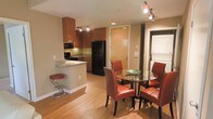 This single-floor unit presents an open-concept layout that integrates the kitchen and dining area into one cohesive living space. The kitchen is outfitted with warm medium-toned wood cabinetry, black appliances including a full-size refrigerator, microwave, and range, and a light granite-topped peninsula that doubles as a breakfast bar with a chrome and red stool. Track lighting runs along the ceiling, providing targeted illumination across both the kitchen and dining areas. The flooring throughout is light hardwood, lending warmth and continuity to the space. The dining area features a round glass-top table surrounded by red upholstered chairs, adding a bold accent against the neutral walls. A stacked washer and dryer unit is visible through an adjacent doorway, offering in-unit laundry convenience. The entry door is centrally positioned, giving the layout a straightforward and functional flow. Overall, the space is compact yet efficiently designed, with consistent finishes and a warm, welcoming ambiance suited for everyday living.