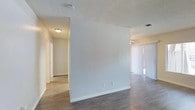 This single-story space offers a straightforward, functional layout designed to maximize usability. The interior features durable wood-look hard flooring throughout, providing a cohesive look that flows seamlessly from room to room. A central dividing wall creates a natural separation between the main living area and the hallway leading to additional rooms, offering a sense of defined zones within an otherwise open floor plan.

The living area is equipped with a ceiling fan, promoting air circulation, while vertical blinds cover what appears to be a sliding glass door, allowing for adjustable natural light and potential access to an outdoor area. The textured popcorn ceiling is consistent throughout the visible spaces, adding a classic residential finish overhead.

The hallway, lit by a flush-mount ceiling light fixture, leads to further rooms and includes multiple doorways, suggesting a practical multi-room configuration. Electrical outlets are conveniently placed along the lower portion of the dividing wall. Overall, the space presents a clean, neutral palette with white walls and light trim, making it a versatile and move-in ready environment.