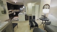 This single-floor residence showcases an open-concept layout that seamlessly connects the kitchen and living area, creating a cohesive and functional living environment. The kitchen is a standout feature, appointed with dark espresso cabinetry that contrasts sharply against light-colored walls and a white kitchen island with granite countertops. Stainless steel appliances add a modern, polished touch, while pendant lighting above the island provides focused task illumination. The island also offers barstool seating, making it ideal for casual dining or entertaining.

Crown molding runs along the ceiling, lending an elegant architectural detail throughout the space. The living area flows naturally from the kitchen, finished with neutral-toned carpet that complements the overall color palette. Accent furniture, including a boldly striped armchair and a sleek side table, add visual interest and character to the space.

A dedicated entryway with a solid white door provides a clear point of entry, flanked by additional doors that suggest closet or utility access. An arched window detail near the entryway allows natural light to filter in. The overall ambiance is contemporary and well-appointed, balancing dark and light contrasts throughout.