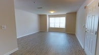 This single-floor space offers a clean, functional layout designed to maximize comfort and usability. The main living area is an open, generously sized room finished with warm hardwood-style flooring that runs throughout, lending a cohesive and inviting feel underfoot. The walls are painted in soft, neutral beige tones that complement the light wood flooring and create a calm, understated ambiance well-suited for a variety of furnishing styles. A large window positioned along one wall serves as a central architectural feature, allowing natural light to flood the interior and providing a connection to the outdoors. Flush ceiling lighting provides additional illumination, and a ceiling-mounted sprinkler reflects the space's attention to practical amenities. Overhead ventilation is provided via a ceiling duct, contributing to the unit's overall comfort. Clean white baseboards frame the perimeter of the room, adding a crisp finishing touch to the overall design. The open floor plan allows for flexible arrangement and makes the space feel airy and unencumbered. Overall, this space presents a straightforward, well-maintained interior with a neutral palette and quality finishes that create a welcoming and versatile environment.