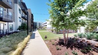 This thoughtfully designed outdoor amenity space serves as a central courtyard within a multi-story residential complex. The grounds are meticulously landscaped, featuring manicured lawns, ornamental grasses, agave plants, mulched garden beds, and young trees that provide shade and visual interest throughout. String lights are strung overhead, adding an inviting ambiance for evening gatherings.

A composite wood deck serves as the focal point of the courtyard, furnished with stylish wicker lounge chairs with green cushions and a decorative gravel feature bed. A pergola with a striped canopy provides shaded seating with additional lounge and sectional furniture, creating a comfortable outdoor retreat. Rows of bamboo-like trees line the perimeter walls, offering a sense of privacy and greenery.

An outdoor kitchen area features a stone-clad grill station equipped with multiple grills, accompanied by dining tables and chairs ideal for al fresco meals. A covered patio with stone columns provides a sheltered transition between indoor and outdoor spaces.

The interior lounge area features tile flooring, tall ceilings with an oversized ceiling fan, white structural columns, and a striking teal textured accent wall. A bar-height dining setup and mounted television make this space suitable for social gatherings. The overall design balances modern aesthetics with comfortable, functional outdoor living.