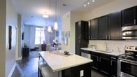 This single-floor residence showcases a thoughtfully designed open-concept layout that seamlessly connects the kitchen and living areas. The kitchen is a standout feature, equipped with rich, dark espresso cabinetry that extends to the ceiling, providing generous storage. A large granite-topped island anchors the space and doubles as a breakfast bar with upholstered seating, making it ideal for casual dining and entertaining. Pendant lighting hangs stylishly above the island, complementing the track lighting installed over the main cabinetry. The countertops feature light-toned speckled granite that contrasts strikingly with the dark cabinetry. Stainless steel appliances, including a dishwasher, and a sleek sink with modern fixtures complete the culinary space. The transition into the living area is smooth and uninterrupted, where neutral-toned walls, hardwood-style flooring, and an elegant chandelier create a warm, sophisticated ambiance. Large windows allow natural light to fill the living space, enhancing the overall sense of openness. The combination of contemporary design elements and functional layout gives this residence a polished, modern character throughout.
