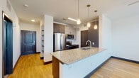 This contemporary single-floor residence is designed with a clean, modern aesthetic throughout. The open-concept kitchen is a standout feature, centered around a large granite-topped island with an undermount sink and pendant lighting hanging above. Taupe cabinetry pairs with a full suite of stainless steel appliances — including a French door refrigerator, microwave, and range — to create a functional and stylish cooking space. The kitchen flows seamlessly into the living and dining area, which is finished with warm hardwood-style flooring, a ceiling fan, and a sliding glass door that invites natural light and outdoor access.

A hallway connects the main living area to the private spaces and features eye-catching built-in open shelving, ideal for display or storage. The bedrooms are carpeted for comfort, each equipped with a ceiling fan and windows fitted with blinds that provide ample natural light while maintaining privacy.

The bathroom continues the granite theme with a matching vanity countertop and dark-framed cabinetry, complemented by a large mirror, a shower-tub combination, and hardwood-style flooring. Throughout the residence, dark door and baseboard trim provides sharp contrast against bright white walls, lending a polished, cohesive look to the entire space.