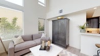 Located at 3851 Harbour Island Ln in Oxnard, CA, this contemporary two-story home offers a well-designed layout with refined finishes and an abundance of natural light. The main living area impresses with dramatically soaring double-height ceilings and large floor-to-ceiling windows that flood the space with daylight, creating an airy, open atmosphere. Light hardwood-style flooring flows throughout the main level, complementing the neutral, modern palette. The dining area sits adjacent to the living room and features direct access to a private outdoor balcony through full-height glass doors, making it ideal for indoor-outdoor living. The kitchen is well-appointed with dark wood cabinetry, white quartz countertops, stainless steel appliances, and ample storage with floor-to-ceiling cabinetry. A sleek powder bath on the main level features a floating vanity, white quartz countertop, and a backlit mirror. Upstairs, the home includes a spacious loft-style area with patterned carpeting and open railings overlooking the lower level, along with a generously sized bedroom complete with plush carpeting and a walk-in closet offering extensive shelving and hanging space. Additional sitting or flex spaces provide versatile options for a home office or lounge. The overall design balances modern architectural detail with comfort and functionality.