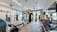 This fitness center is a thoughtfully designed, single-floor workout facility offering a spacious and well-organized environment for a full range of exercise activities. The interior is finished with neutral gray walls and a checkered carpet tile floor that adds a modern aesthetic while providing comfort and durability underfoot. Ceiling fans and linear LED lighting are installed throughout, ensuring excellent ventilation and bright, even illumination across the entire space.

The facility is lined with large windows along multiple walls, flooding the interior with abundant natural light and providing views of the surrounding outdoor greenery. A wall of mirrors further enhances the sense of openness and allows users to monitor their form during exercise.

The cardio zone features multiple treadmills and elliptical machines, each equipped with individual display screens. The strength training area is extensively equipped, including a multi-functional cable and pulley rack system, a Smith machine, a dedicated punching bag station with TRX suspension straps, a weight bench, a full dumbbell rack, kettlebells, medicine balls, and a leg extension machine. Built-in shelving provides organized storage for accessories and equipment. The facility also includes a flat-screen television mounted on the wall. Entry is through glass-paneled doors, and posted fitness center guidelines are visible near the entrance.