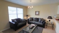This single-floor residence offers a comfortable and well-appointed living environment designed for both relaxation and everyday functionality. The living room serves as the heart of the space, centered around a stylish multi-arm chandelier that adds a touch of elegance to the room. Warm, neutral-toned walls create a calm and inviting ambiance throughout. The seating arrangement features a generously sized sofa and loveseat in a combination of dark fabric and leather upholstery, complemented by plush accent pillows. A glass-topped coffee table with a metal frame anchors the seating area, adding a sleek, modern contrast to the softer furnishings. A textured area rug grounds the arrangement and adds warmth underfoot. Natural light pours in through a large window dressed with horizontal blinds, brightening the room and offering a view of the surrounding greenery outside. A classic table lamp provides additional ambient lighting for the evenings. Wall art and thoughtful décor touches lend a personalized character to the space. The carpeted flooring runs throughout, contributing to the cozy, lived-in feel of this single-level home.