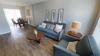 This single-floor space offers a cohesive and inviting layout designed for comfort and functionality. The living area serves as the centerpiece, showcasing warm hardwood-style flooring that runs throughout and lends a sense of continuity to the space. Neutral gray walls provide a clean, contemporary backdrop that complements the carefully selected furnishings.

The seating area is anchored by a plush blue-gray sofa adorned with a tasteful mix of decorative pillows, paired with a matching accent chair that creates a comfortable conversational arrangement. A round wooden coffee table with a distinctive crossed-leg base adds a mid-century modern touch, while coordinating side tables with warm-toned lamps contribute to the ambient lighting of the room.

Wall décor features a series of framed botanical prints arranged vertically, adding an artistic focal point that ties the room's natural tones together. Additional artwork is visible along the adjacent wall, reinforcing a curated, gallery-like aesthetic throughout the space.

The open layout allows for easy flow between areas, with a glimpse into a neighboring room suggesting a well-connected floor plan. Overall, the space balances modern design sensibilities with approachable warmth, making it both visually appealing and highly functional.
