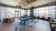 Located at 4850 Rugby Avenue in Bethesda, Maryland, this upscale amenity lounge is designed with a contemporary aesthetic that blends rich materials, layered lighting, and expansive floor-to-ceiling windows that frame sweeping views of the surrounding cityscape. The open layout flows seamlessly between distinct zones, each thoughtfully curated for both social and private use. A striking game area anchors the space with a billiard table set atop a vibrant geometric area rug, illuminated by sculptural branched light fixtures and bordered by glass doors leading to an outdoor terrace. Adjacent to the game room is a comfortable lounge with a large-screen television, tufted leather sofa, and eclectic accent furnishings set against a reclaimed wood feature wall. The bar area showcases a dramatic marble accent wall, dark wood cabinetry, a long concrete countertop with integrated sink, and decorative tile backsplash details. A dining and meeting zone features a combination of upholstered booth seating with textured tile surrounds, open communal tables with leather and mixed-fabric chairs, and a wall-mounted display screen. Warm wood-look flooring in a herringbone pattern, industrial-style Edison bulb track lighting, and glass-paneled sliding doors with frosted accents complete the refined, urban atmosphere throughout.