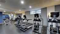 The fitness center at 1303 Delaware Ave in Wilmington, Delaware is a dedicated single-floor exercise space designed to support an active lifestyle. The room features a drop ceiling with recessed lighting that evenly illuminates the space, complemented by a horizontal window along one wall that allows natural light to filter in. The flooring consists of durable rubber gym matting, providing both safety and comfort during workouts. A large wall mirror expands the sense of space and allows users to monitor their form during exercise. The facility is equipped with True-brand cardio machines, including multiple treadmills and an elliptical trainer, reflecting a commitment to quality fitness equipment. Additional strength and conditioning equipment is visible toward the rear of the space. The overall aesthetic is clean and functional, with a neutral color palette of grays and whites contributing to a focused workout environment. The layout is thoughtfully arranged to maximize usable floor space while maintaining comfortable circulation between equipment. This well-maintained fitness center offers residents a convenient and professional-grade exercise facility on-site.